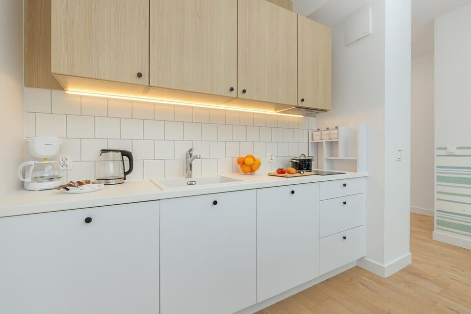 A modern kitchen features white lower cabinets with black handles and light wood upper cabinets, illuminated by a strip of warm light underneath.