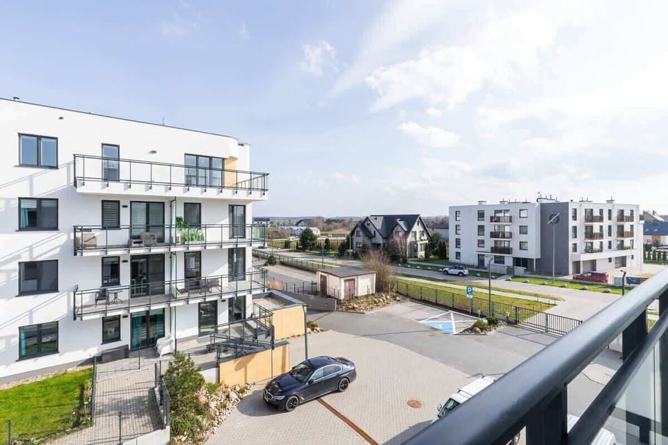 The flat's balcony overlooks a modern residential area with white buildings and balconies, while the green landscape and fragments of urban development can be seen in the background.