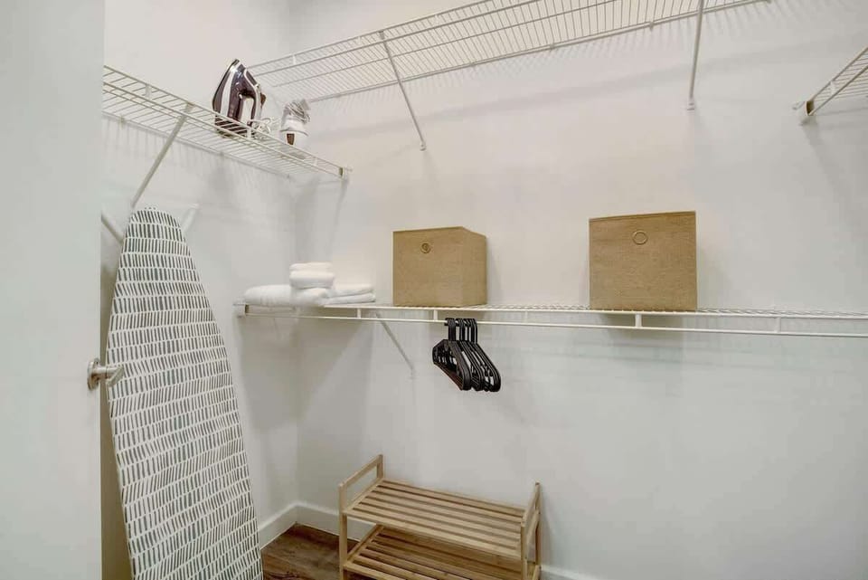 A walk-in-closet with plenty of shelves, hangers, iron & board, shoe rack, crib, high chair, and vacuum
