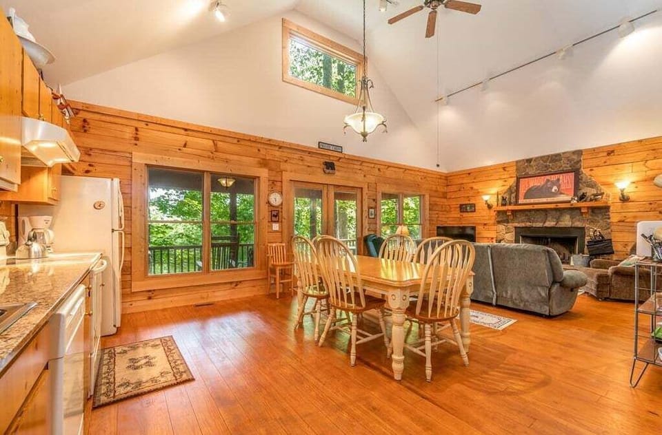 Open-concept interior with dining table, living room seating, vaulted ceiling, and large windows overlooking the trees. Natural wood finishes create a warm mountain cabin atmosphere.