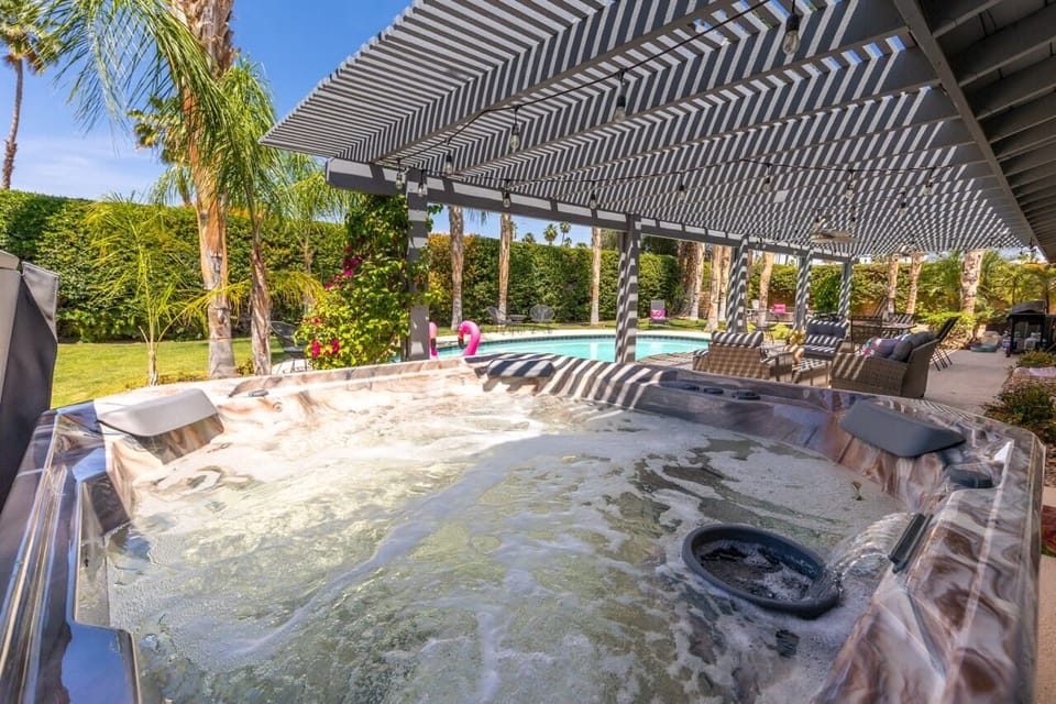 Bubbling hot tub under louvered pergola with pool and palm garden views — soak while the flamingos float