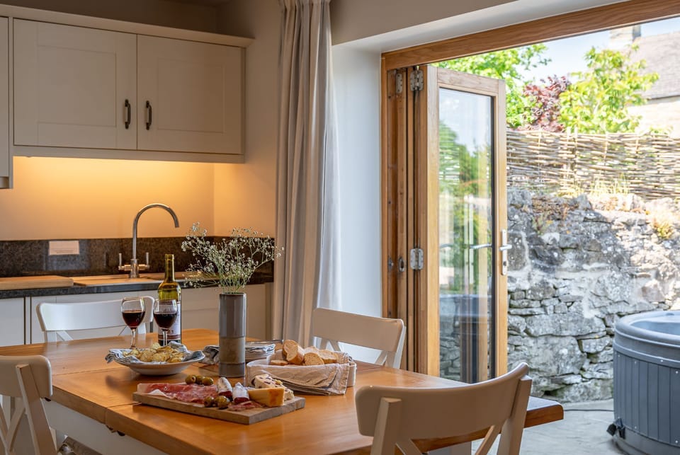 Dining area at Woodcroft Cottage with wooden table, locally inspired food setup and patio doors leading to hot tub.