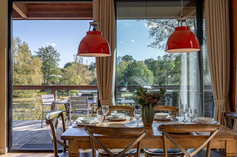 Stylish dining space at Imogen lodge at Brompton Lakes with large windows with lakeside views, wooden table, and red pendant lights.