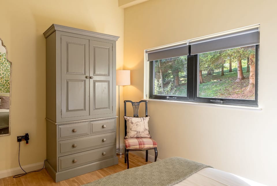 Bedroom in Imogen lodge at Brompton Lakes with wardrobe, chair, and window overlooking trees and garden.