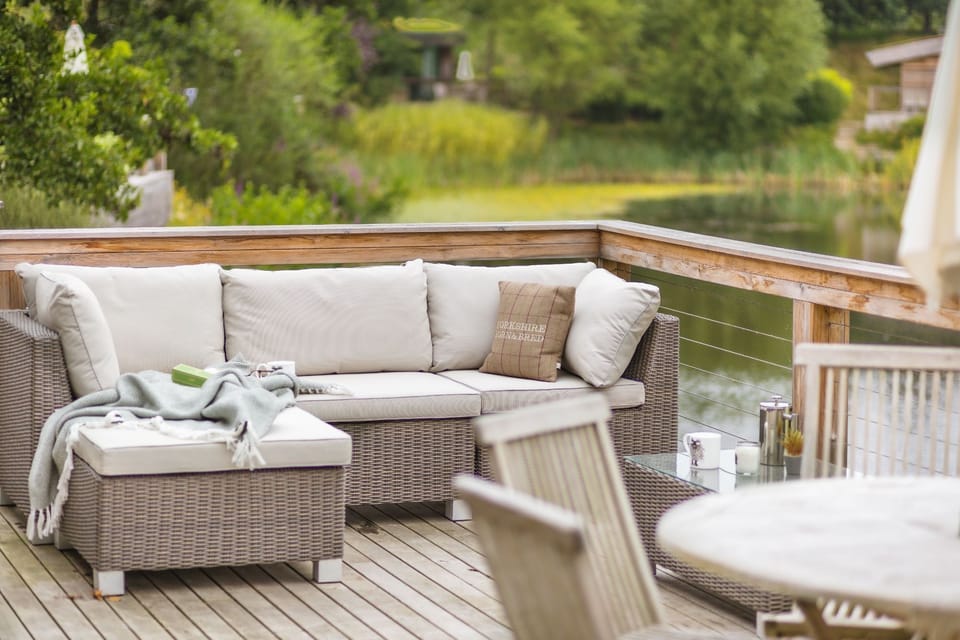 Outdoor decking area with wicker sofa seating, cushions, a small table with cups, and a view over a lake and greenery.