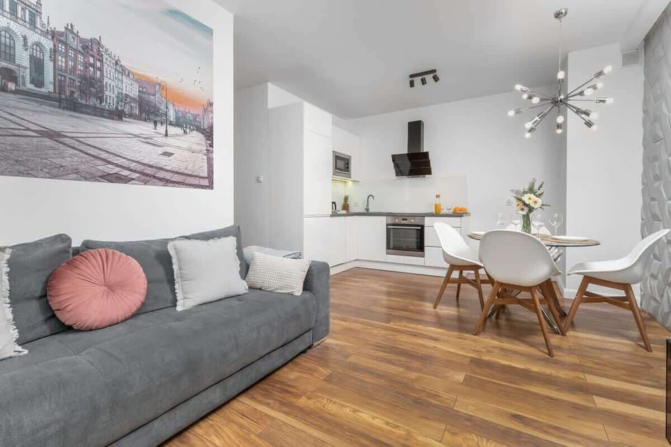 A bright, modern living room with a grey sofa, wooden flooring, large windows, and a wall-mounted TV.