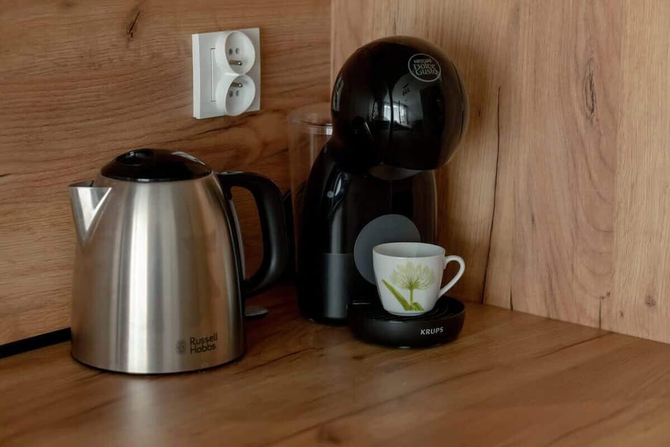 A wooden kitchen shelf with an electric kettle, cup, and a lamp creating a warm beverage station.