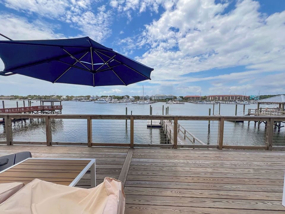 A private waterfront deck featuring sunbeds and a blue umbrella overlooks a tranquil marina with docked boats and distant coastal buildings framed by a scenic sky.