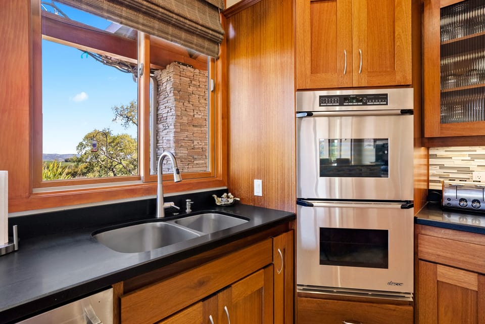 This sophisticated culinary space features high-end stainless steel double ovens integrated into custom wood cabinetry. A modern undermount sink sits beneath a window, offering serene hillside views.