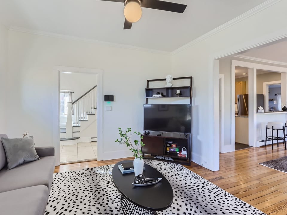 Modern living room with a gray sofa, black coffee table, and a TV on a stand. A ceiling fan is on, and in the background, there are open doorways to a kitchen and a staircase. A plant adorns the table.