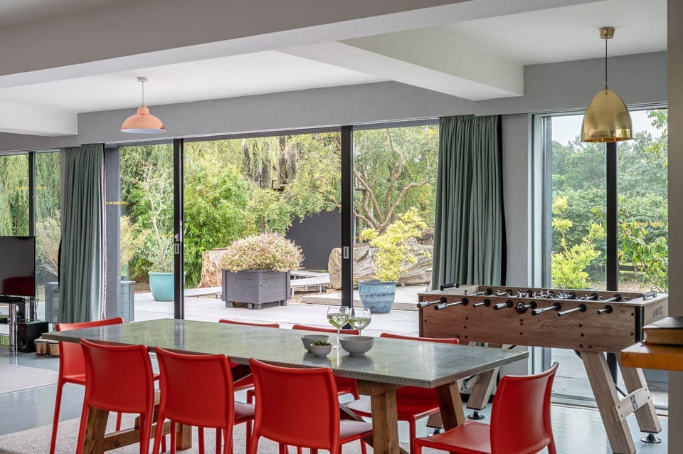 Dining area with large table and red chairs beside a foosball table, with floor-to-ceiling glass doors opening onto a leafy garden terrace.