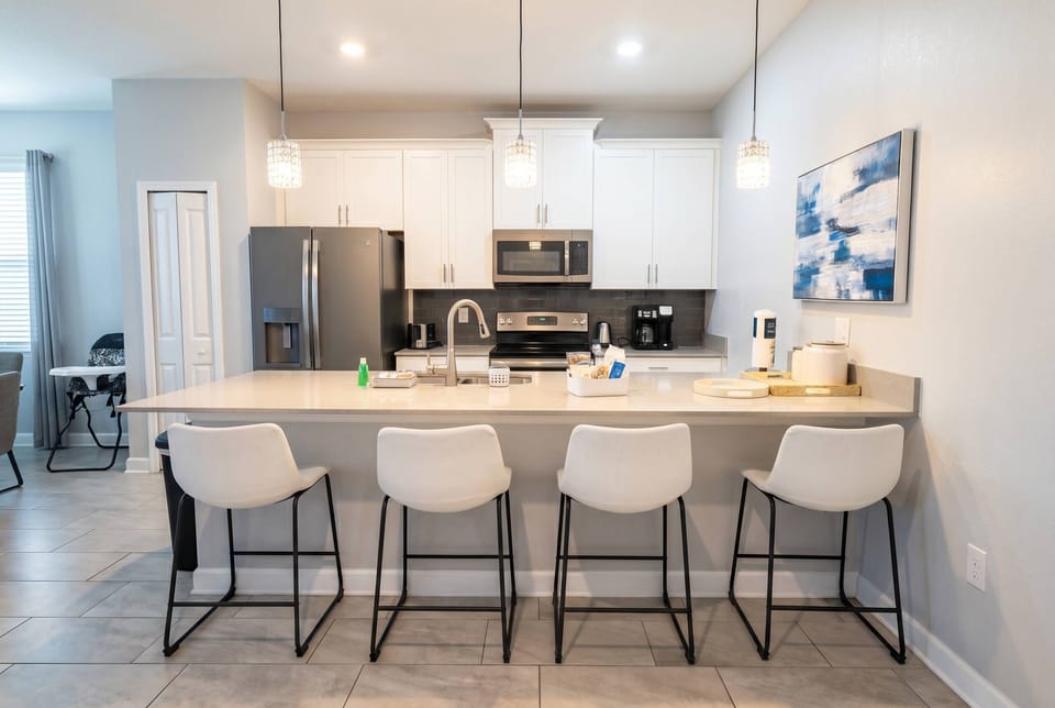 This modern kitchen boasts sleek white cabinetry and stylish gray tile flooring, creating a bright, inviting space. The spacious island with chic bar stools and pendant lighting is ideal for casual dining and gatherings.