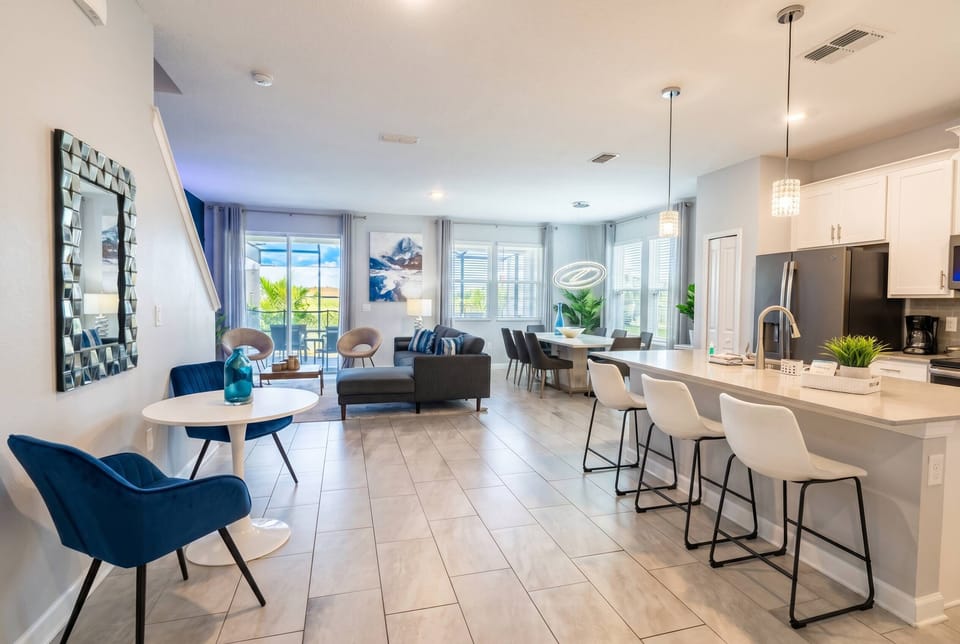 Experience a bright and airy living area with sleek tile flooring and large windows adorned with soft curtains. Modern furnishings, including stylish accent chairs and a chic dining setup, invite relaxation and enjoyable.