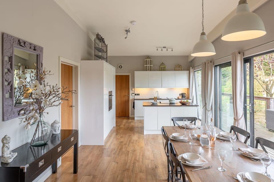 Open-plan kitchen and dining area at Abbey, with a central island, dining table for six, and large doors leading to the lakeside deck.