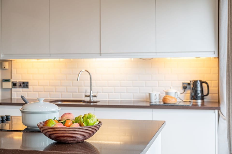 Kitchen at Abbey, with white cabinets, tiled backsplash, sink, kettle on the worktop, and a kitchen island with a bowl of fruit.