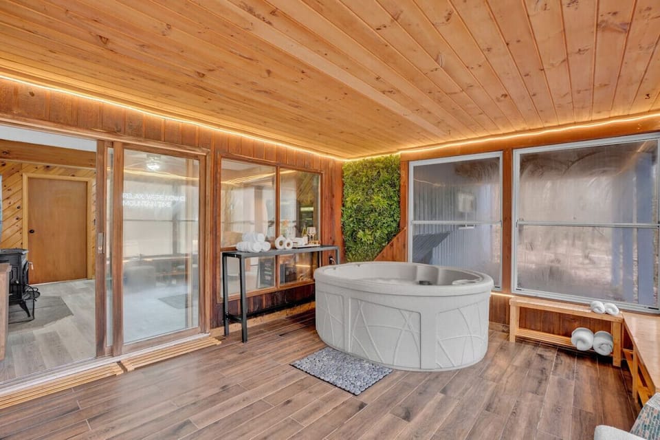 Inviting sunroom with a private hot tub, wood-paneled walls and ceiling, and large windows that let in natural light. Includes a small table for towels and accessories, creating a perfect spot for relaxation.