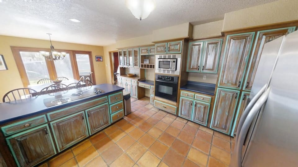 Step into your spacious kitchen with rustic turquoise cabinetry, modern appliances, and terracotta tile floors. Gather around the island or dine at the adjoining table bathed in natural light from the patio doors.