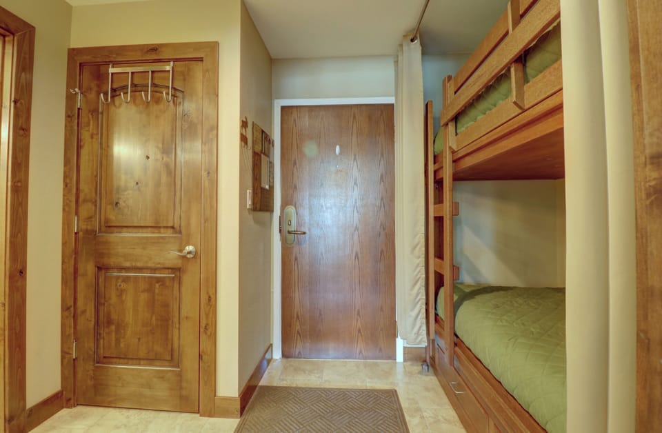 Entryway with a wooden door, beige walls, and a bunk bed to the right with green bedding. A closed door is on the left, and the floor is tiled in beige. A brown mat lies in front of the main door.