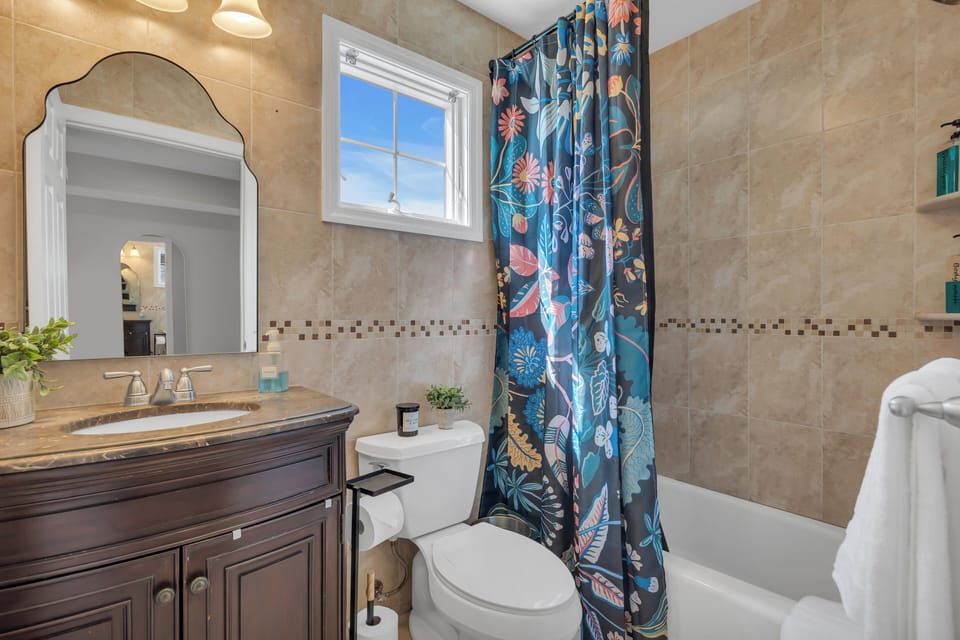 Full bath with travertine tub-shower combo, dark wood vanity with granite top, arched mirror, floral shower curtain, and decorative mosaic tile accent stripe.