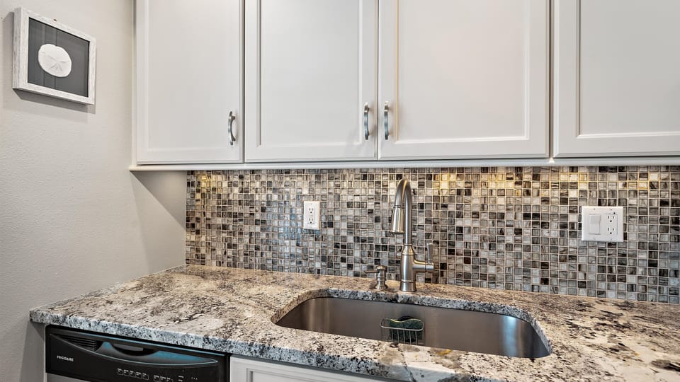 A kitchen sink with a granite countertop beneath white cabinets and a mosaic tile backsplash.