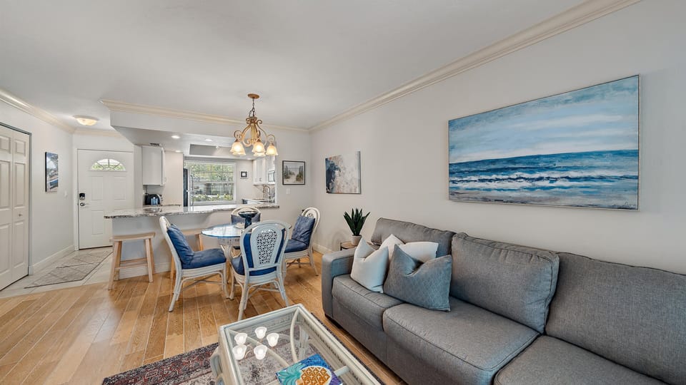 A cozy living and dining area with a grey sofa, blue and white cushions, a glass coffee table, a dining table with four chairs, and ocean-themed artwork on the walls. A kitchen is visible in the background.