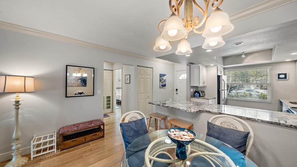 A modern kitchen and dining area featuring a round glass dining table with blue velvet chairs, granite kitchen countertops, and contemporary lighting. The room has light grey walls and wood-look flooring.