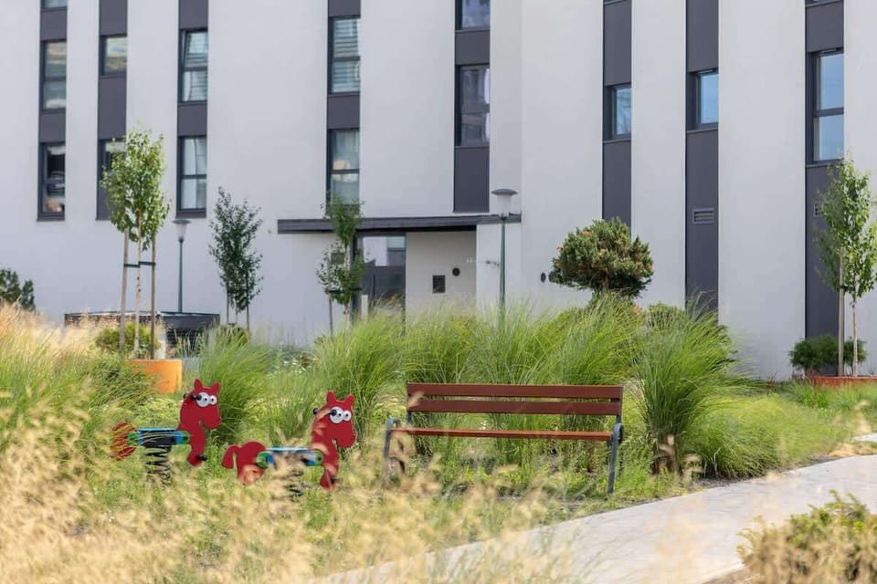 A landscaped garden area with walking paths, benches, and young greenery surrounding the building.

