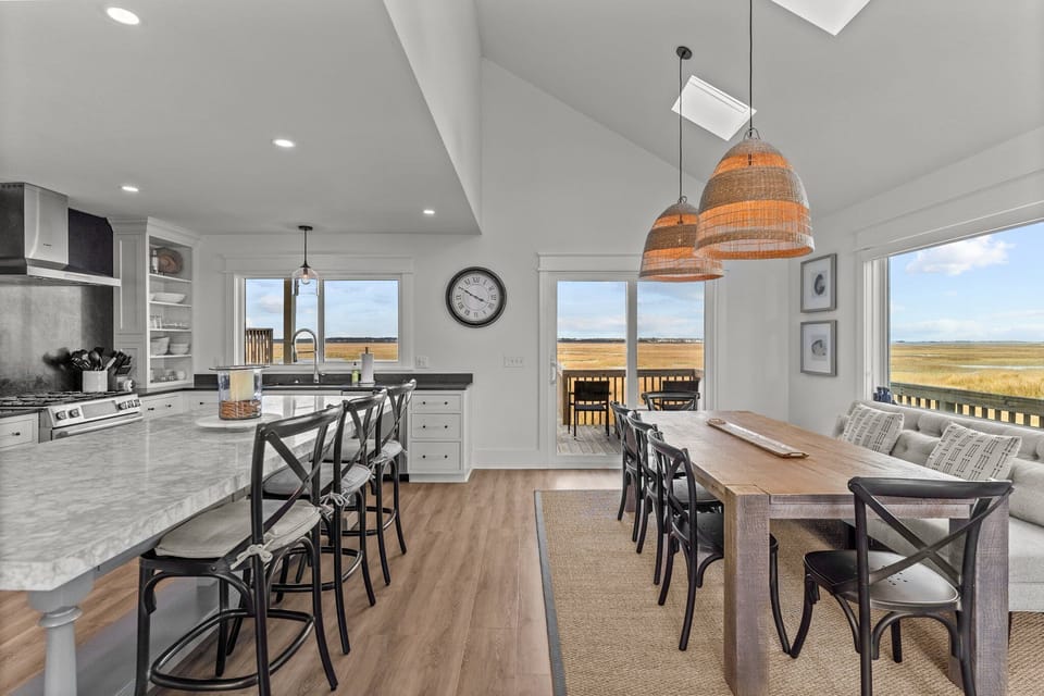 Open dining and living space with a large table, woven pendant lights, fireplace, vaulted ceilings, tall windows, French doors, and bright coastal seating.