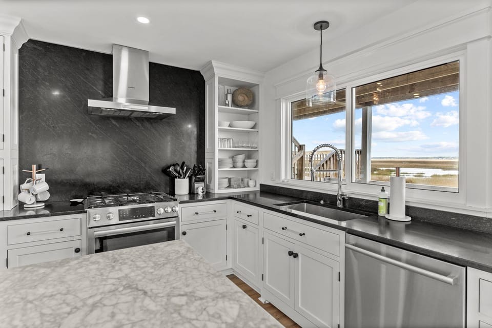 Bright kitchen with a large island, barstools, black countertops, white cabinets, open shelves, stainless range hood, farmhouse sink, and wood-look floors.