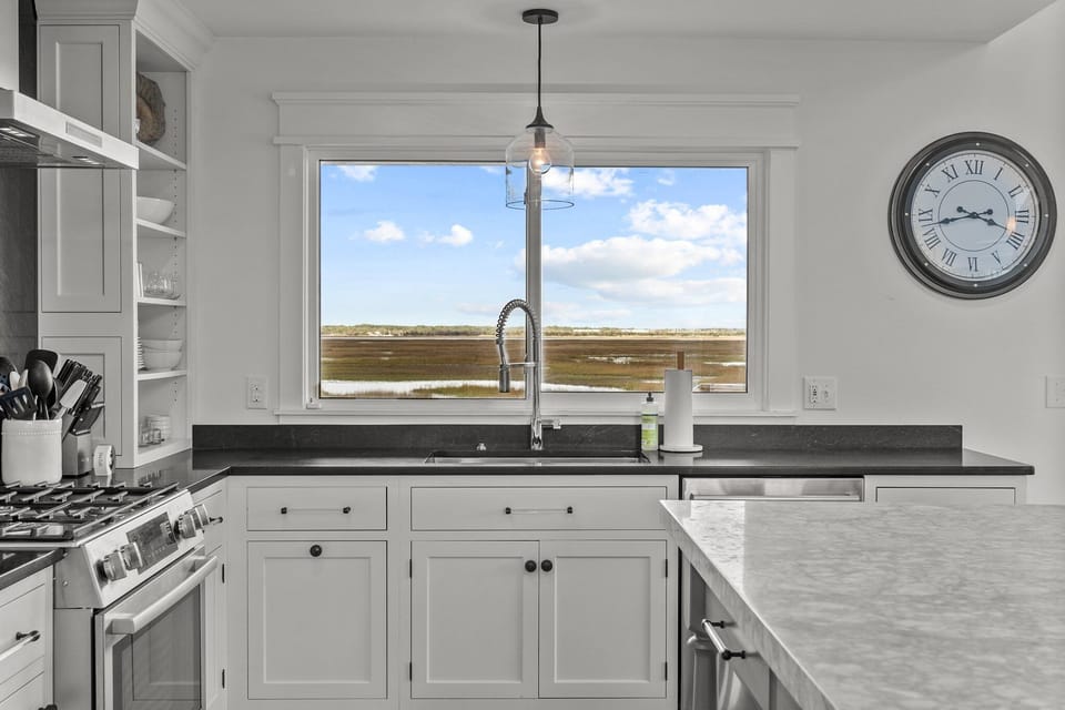 Bright kitchen with a large island, barstools, black countertops, white cabinets, open shelves, stainless range hood, farmhouse sink, and wood-look floors.