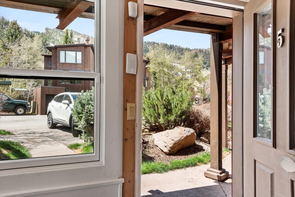 View from inside a house looking out through an open door to a porch, with a white car parked outside, surrounding greenery, and neighboring houses in the background.