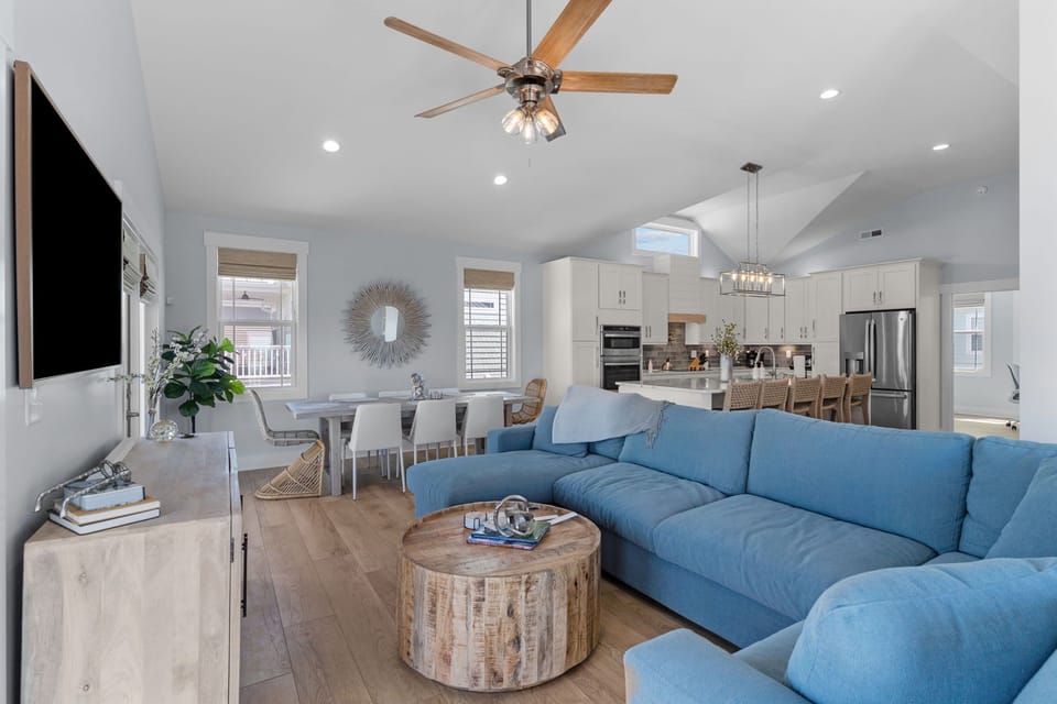 Bright living room with blue sectional, wall-mounted TV, wood console, round coffee table, ceiling fan, balcony doors, ocean glimpses, and wood floors.