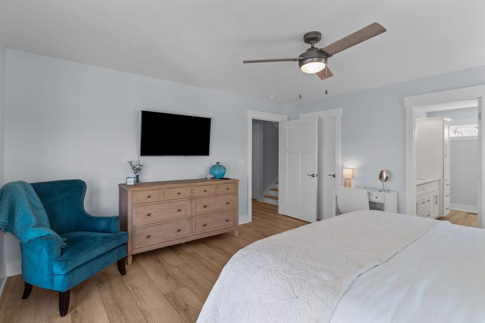 Oak Island bedroom with white bed, ceiling fan, TV, wood dresser, blue chair, balcony doors, wood floors, and open closet with hooks and baskets.