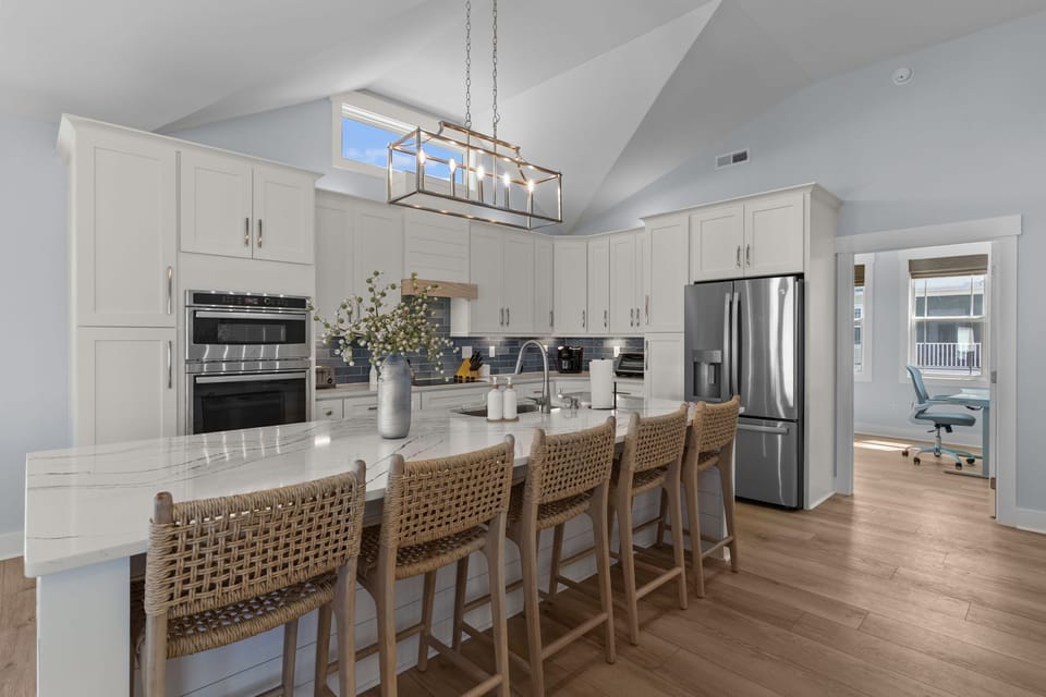 Bright kitchen with vaulted ceiling, large island, woven stools, white cabinets, blue tile backsplash, stainless appliances, modern chandelier, and office nook.