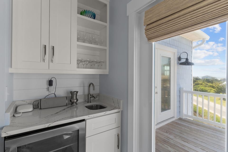 Oak Island living area with wet bar, mini fridge, white cabinets, glass doors, woven shades, balcony access, wood floors, blue sofa, and distant ocean views.