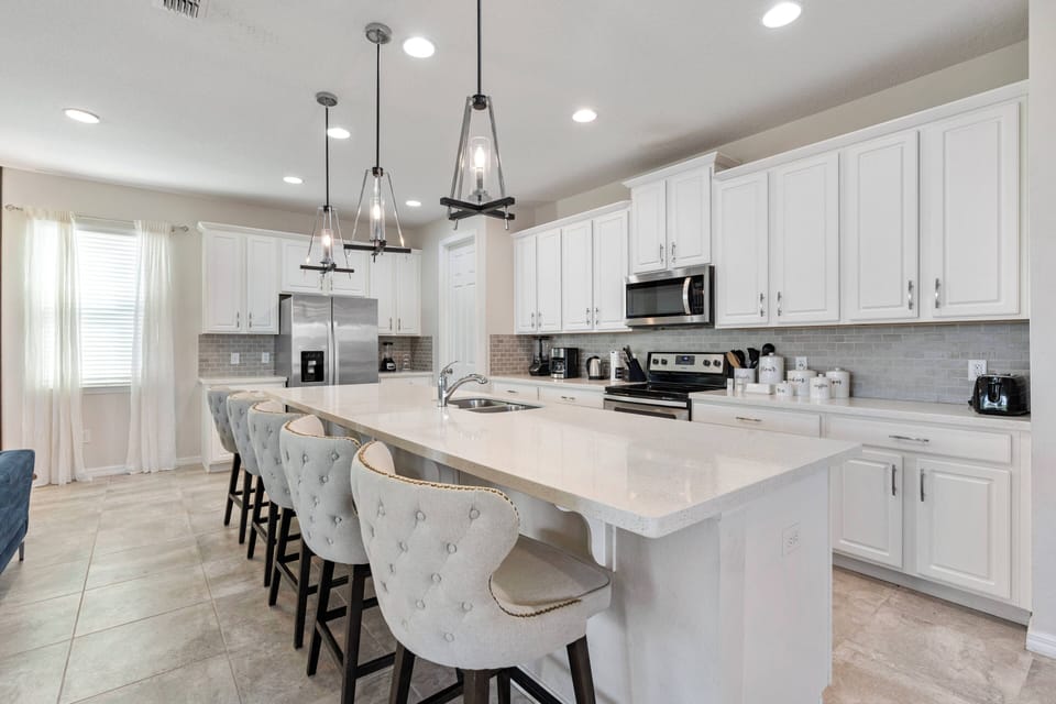 Experience the essence of your vacation in this beautiful kitchen, featuring a spacious island with cozy bar stools and bright white cabinetry, all bathed in warm natural light from large windows, perfect for culinary adventures.