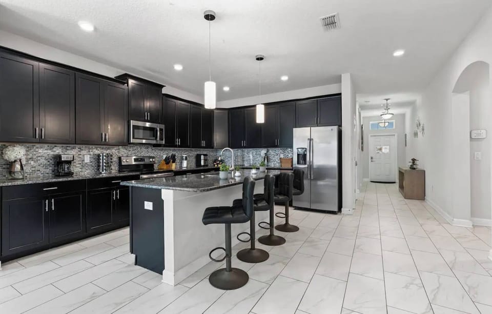 Welcome to this stylish kitchen with sleek black cabinets and light tile flooring. The spacious island with modern bar stools invites casual dining and conversation, all under bright lighting that creates a warm atmosphere.