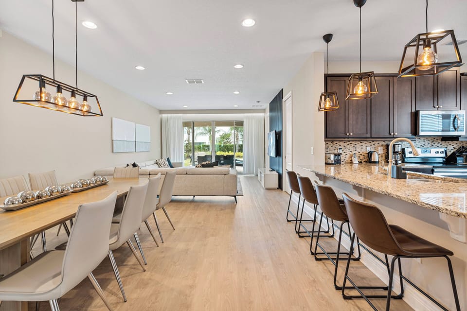 Enjoy the heart of your vacation in a spacious dining area featuring a stylish wood table and modern chairs. Warm pendant lights illuminate the open layout, seamlessly connecting to a cozy living space with inviting views.