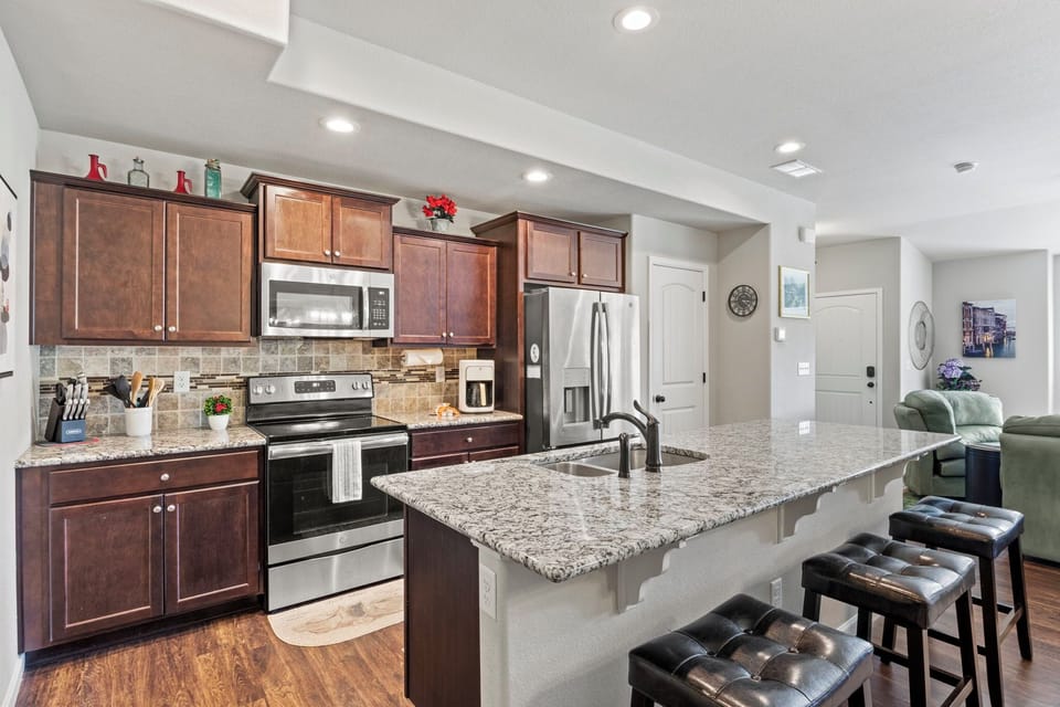 Modern kitchen with dark wooden cabinets, stainless steel appliances, granite countertops, and a large island with three leather stools. Open layout with a living area in the background.