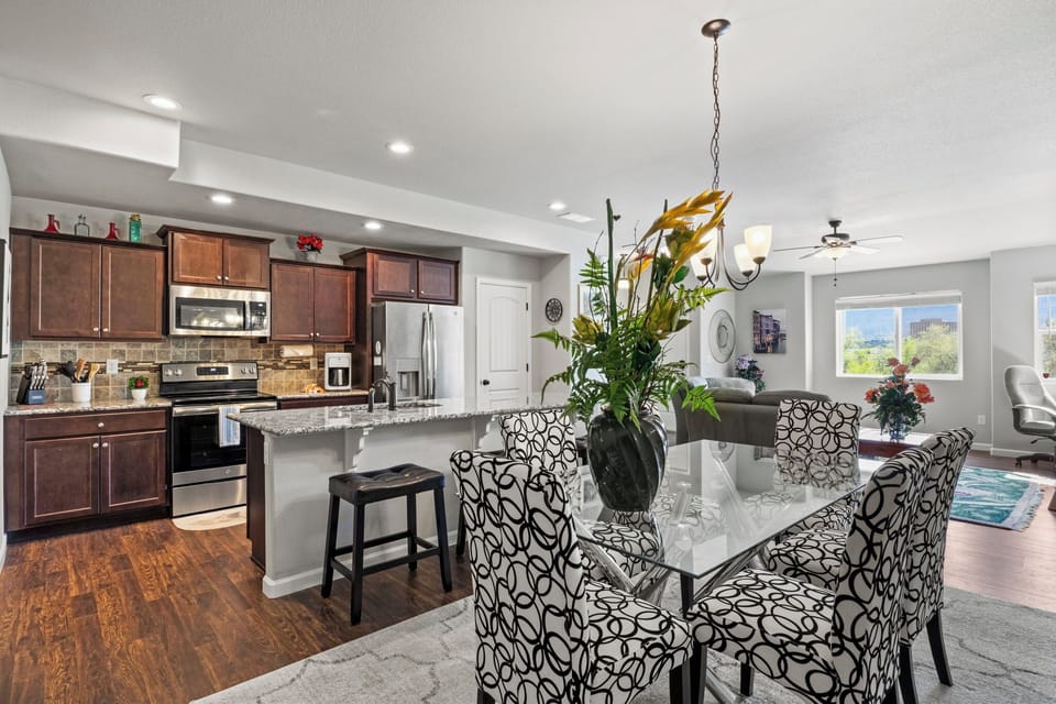 A modern kitchen and dining area featuring dark wood cabinets, a marble countertop island, a glass dining table with patterned chairs, and a large potted plant as the centerpiece.