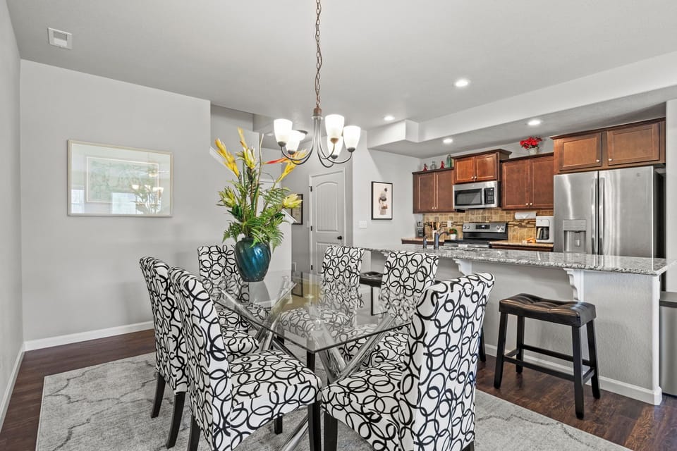 A dining area with a glass-top table, six patterned chairs, and a flower vase centerpiece, adjacent to a kitchen with wooden cabinets, stainless steel appliances, and a breakfast bar.
