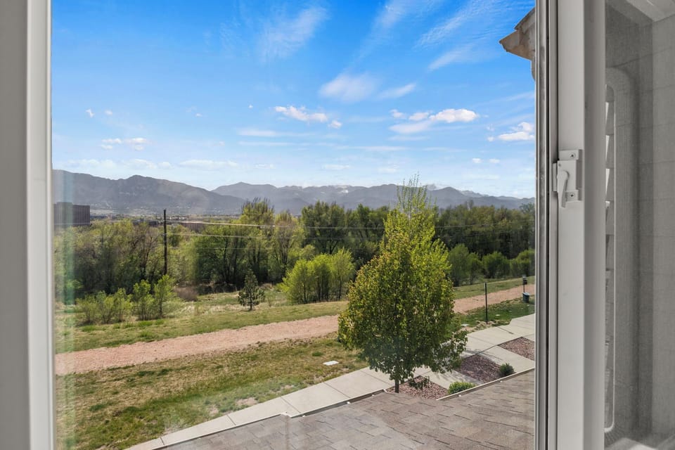 View of mountains and blue sky from a window, overlooking a pathway, grass, and trees in the foreground.