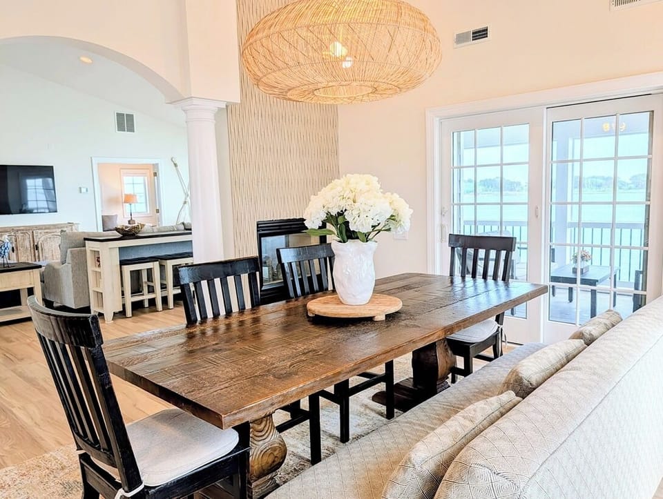 33 Inlet View North Bethany Beach Dining Room - A dining area featuring a rustic wooden table, dark chairs, and a bench, illuminated by a large woven pendant light, with views of the outdoors through sliding glass doors.