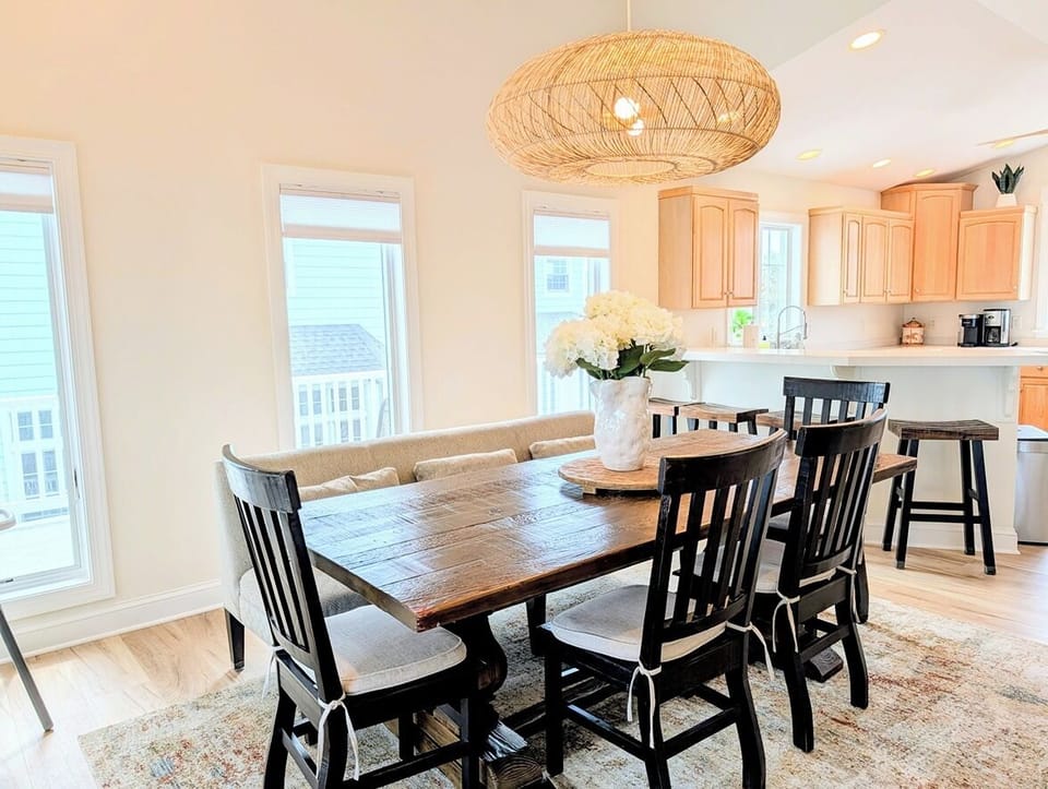 33 Inlet View North Bethany Beach Dining Area - A dining area featuring a rustic wooden table, dark chairs, a bench, and a woven pendant light, with a kitchen visible in the background.