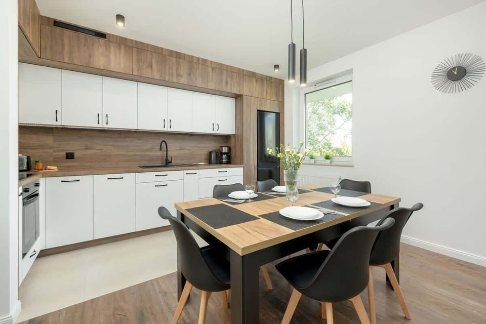 A modern kitchen with white cabinets and wooden accents, a black sink, and a full set of appliances adjoins a dining area with a wooden table, black chairs, and elegant tableware, all illuminated by a large window letting in natural light.