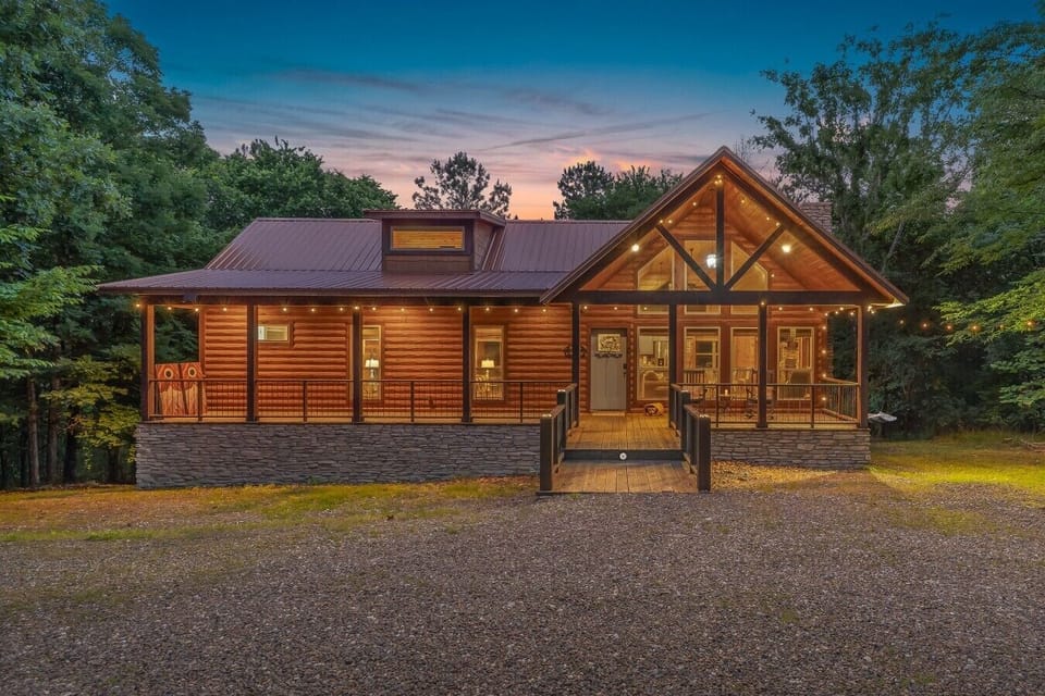 Twilight Pines illuminated at dusk — a stunning A-frame cabin with string lights, stone foundation, and forest views.