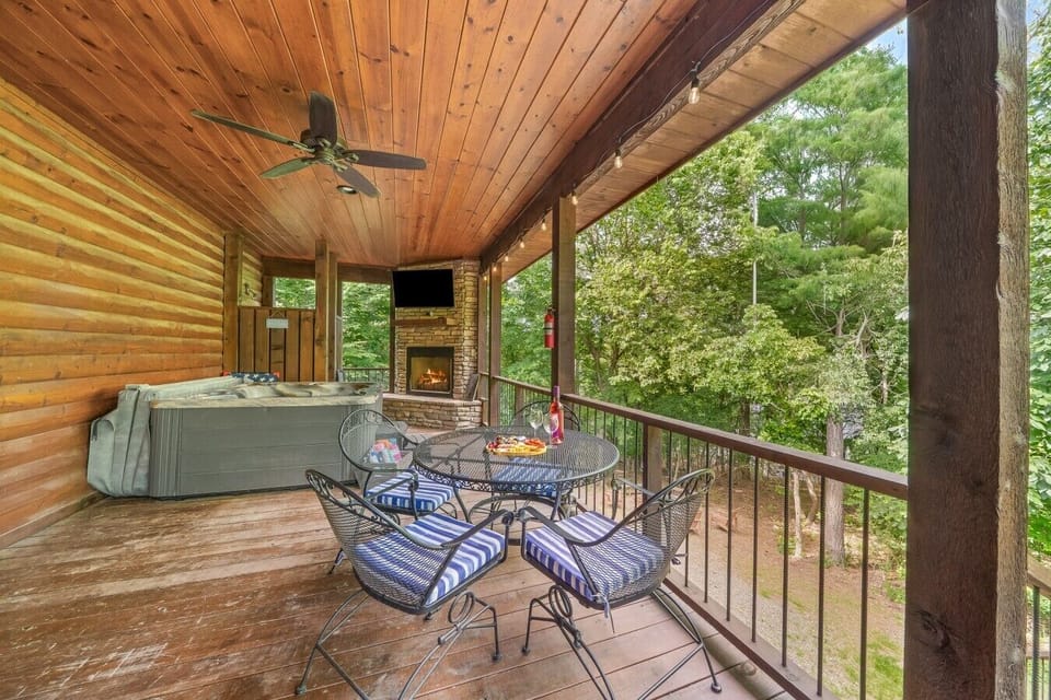 Covered deck featuring private hot tub alongside a stone wood-burning fireplace and mounted TV.