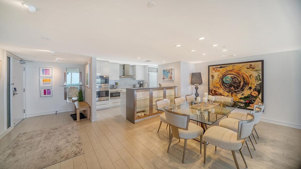 Glass dining table seats eight with cushioned chairs, set against light wood flooring and a large abstract painting.