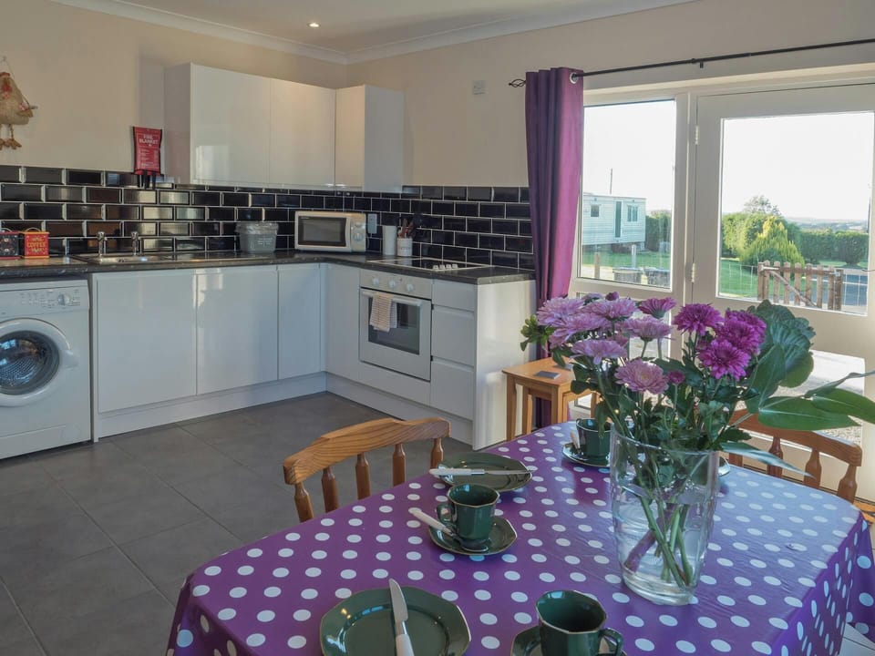 Table laid for tea, bunch of flowers and L-shaped kitchen area with washing machine, built-in oven and hob and microwave