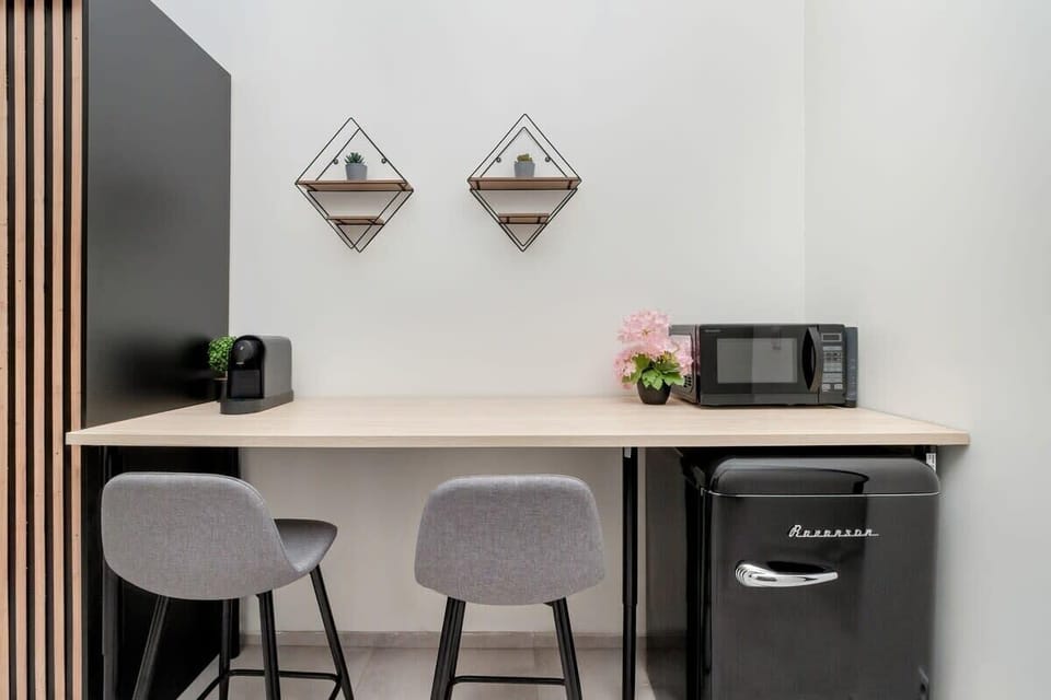 A compact dining nook with a small table, two stools, and geometric wall shelves above a black coffee station.

